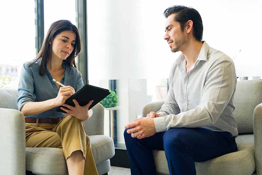 Two people talk on a couch, one holding a tablet.