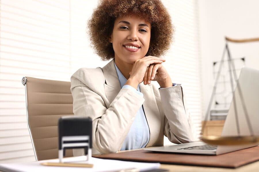 Smiling female Notary sitting at a desk with a laptop and notary stamp.